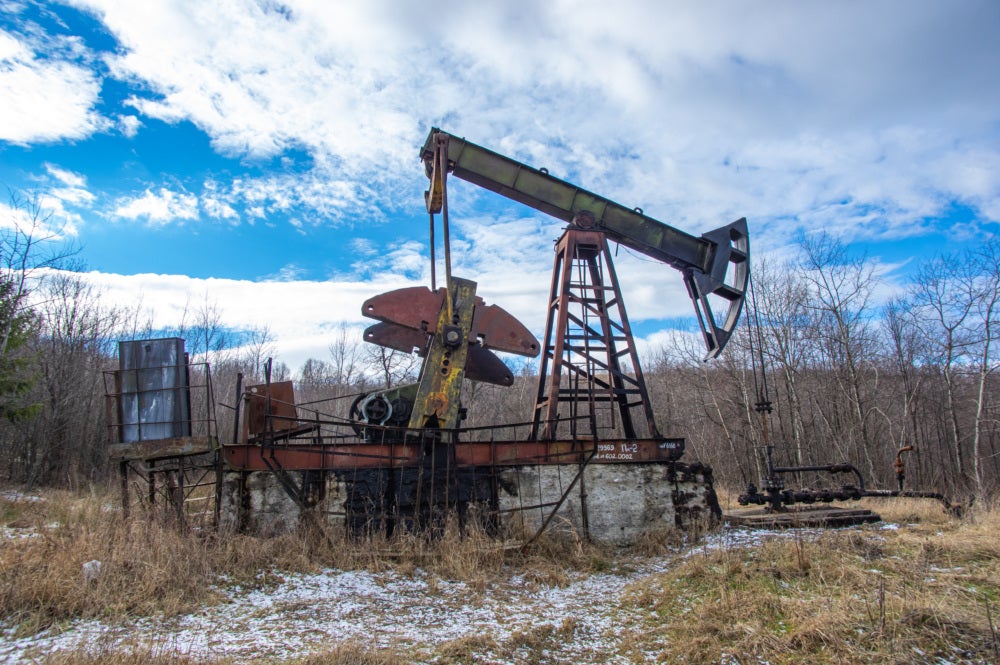 An abandoned oil well is rusting on a brown field. There is a bright blue sky with white clouds and bare trees in the background.
