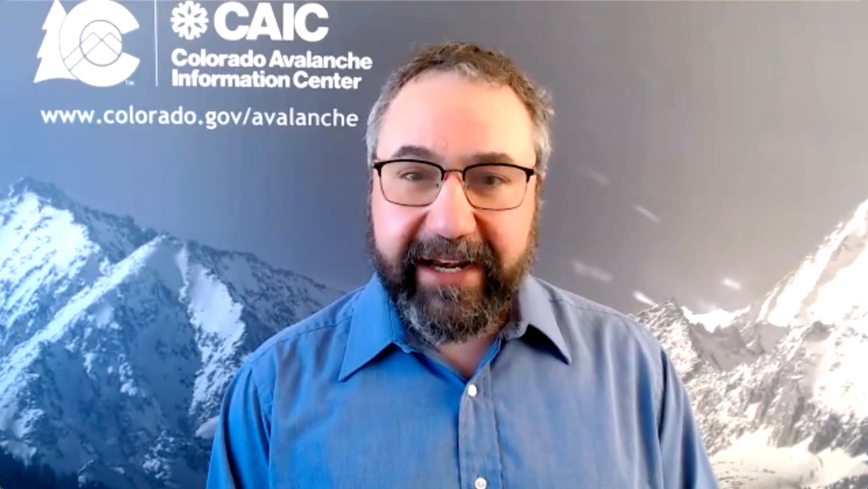 Dr. Ethan Greene is wearing a blue dress shirt and has dark glasses and a dark beard. He is in front of a snow capped mountain background speaking about avalanches.